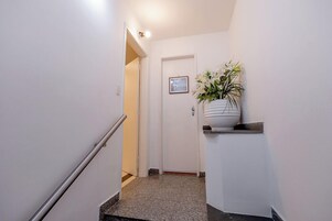 A narrow hallway with a white door at the end, a framed picture on the wall, and a potted plant on a shelf.