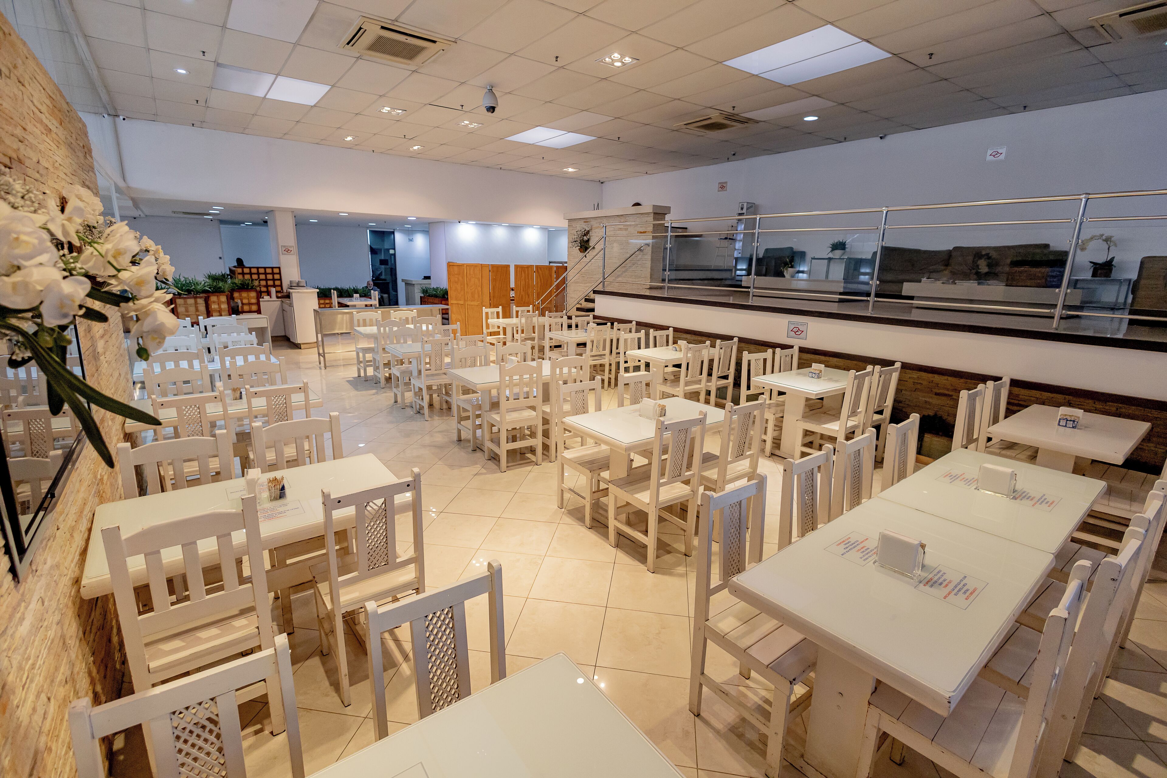 Un restaurant spacieux avec des tables et des chaises blanches, un mur de briques sur un côté et un escalier menant à un niveau supérieur.