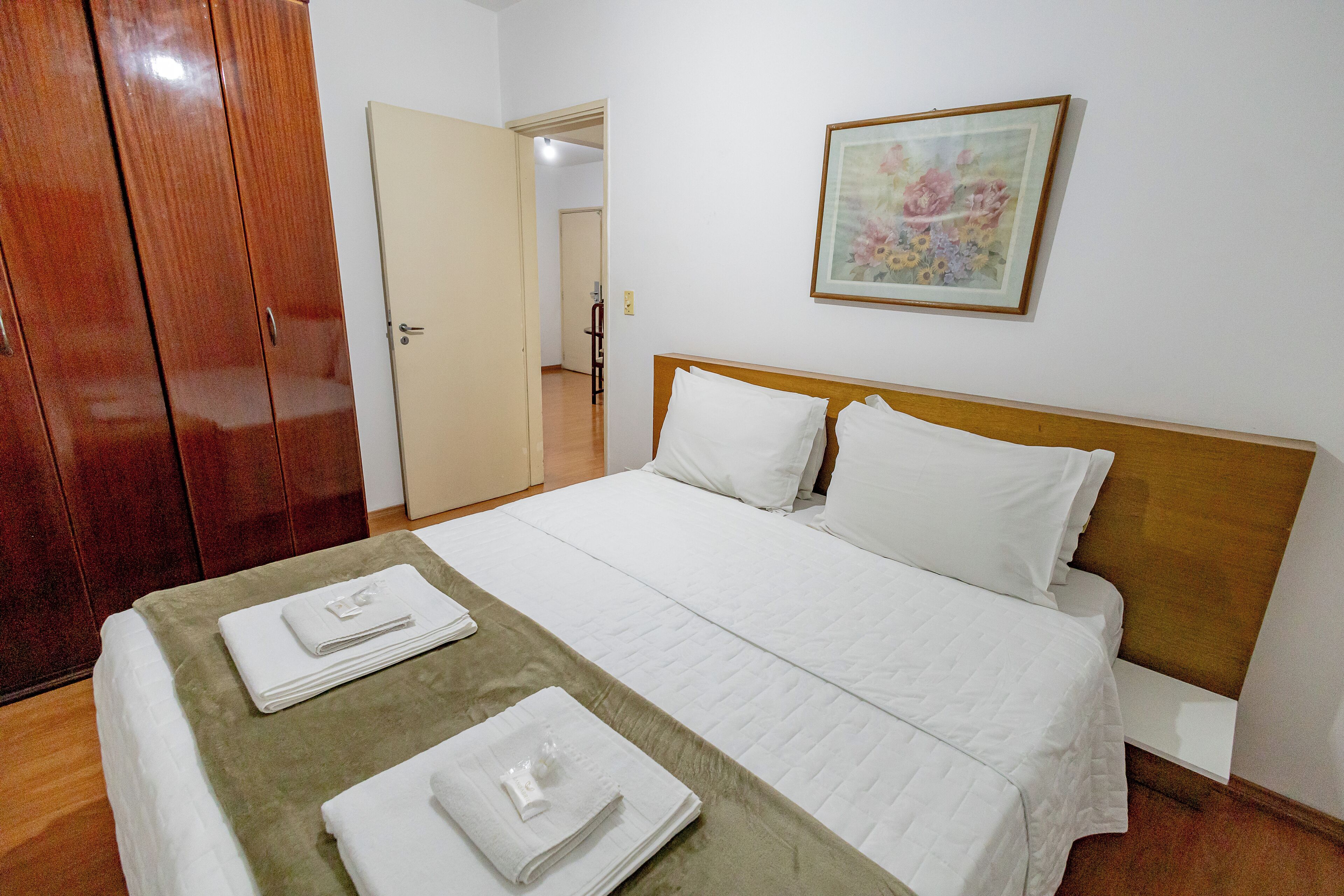 Una cama bien tendida con sábanas blancas, cabecera de madera y colcha verde. A la izquierda se encuentra un armario de madera y una puerta que da a otra habitación. Encima de la cama, hay un cuadro con motivos florales.