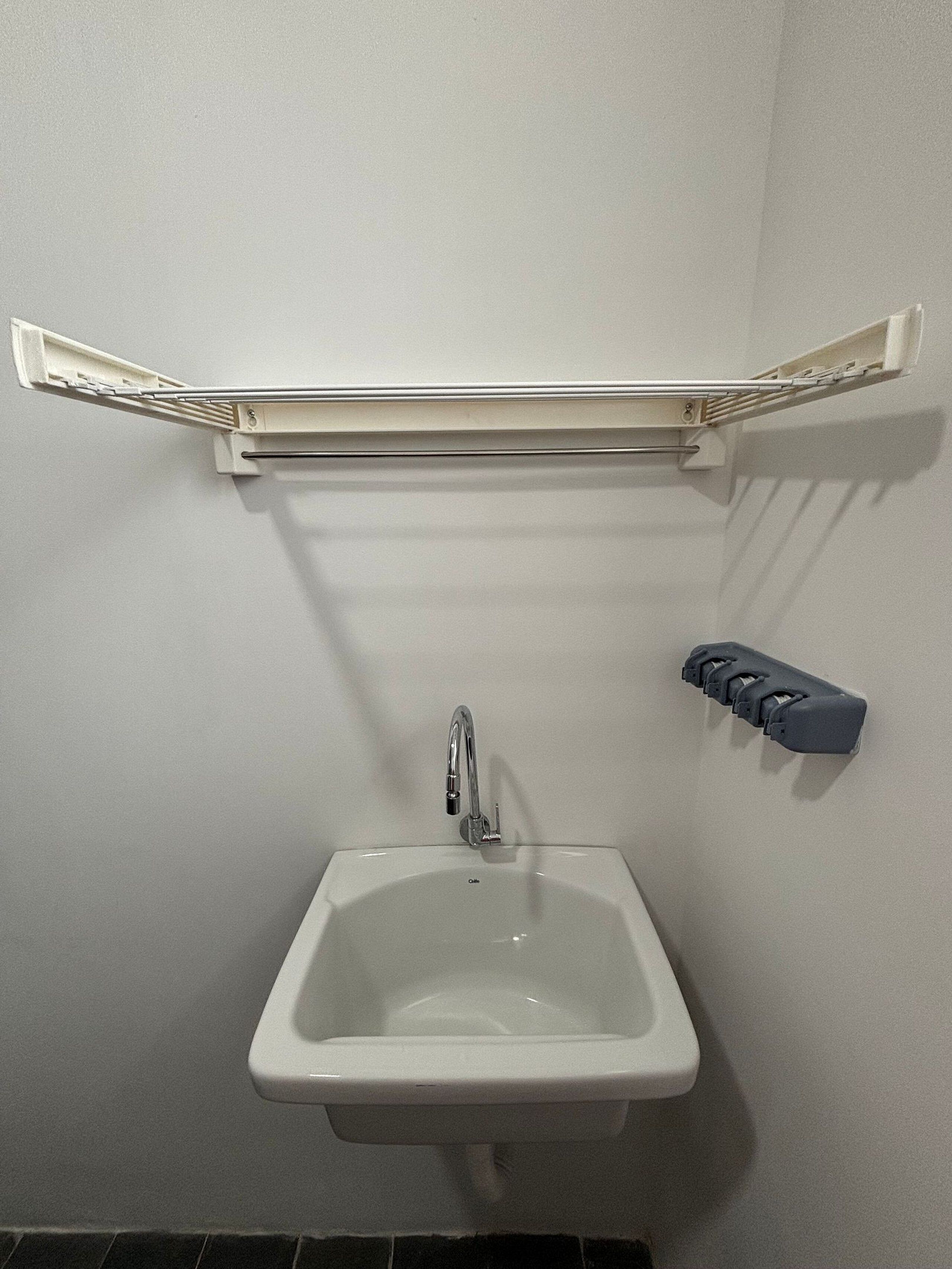 Un lavabo blanc dans la salle de bain, avec un robinet chromé, un porte-serviettes blanc au-dessus et un ensemble de distributeurs de savon gris sur le côté droit.