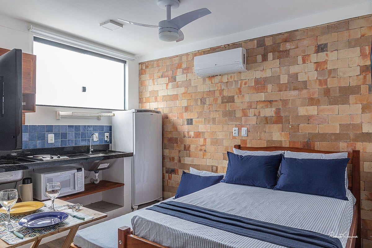 Ein kompakter Raum mit einem Bett, einer kleinen Küchenzeile, einem Fenster und einem Deckenventilator.