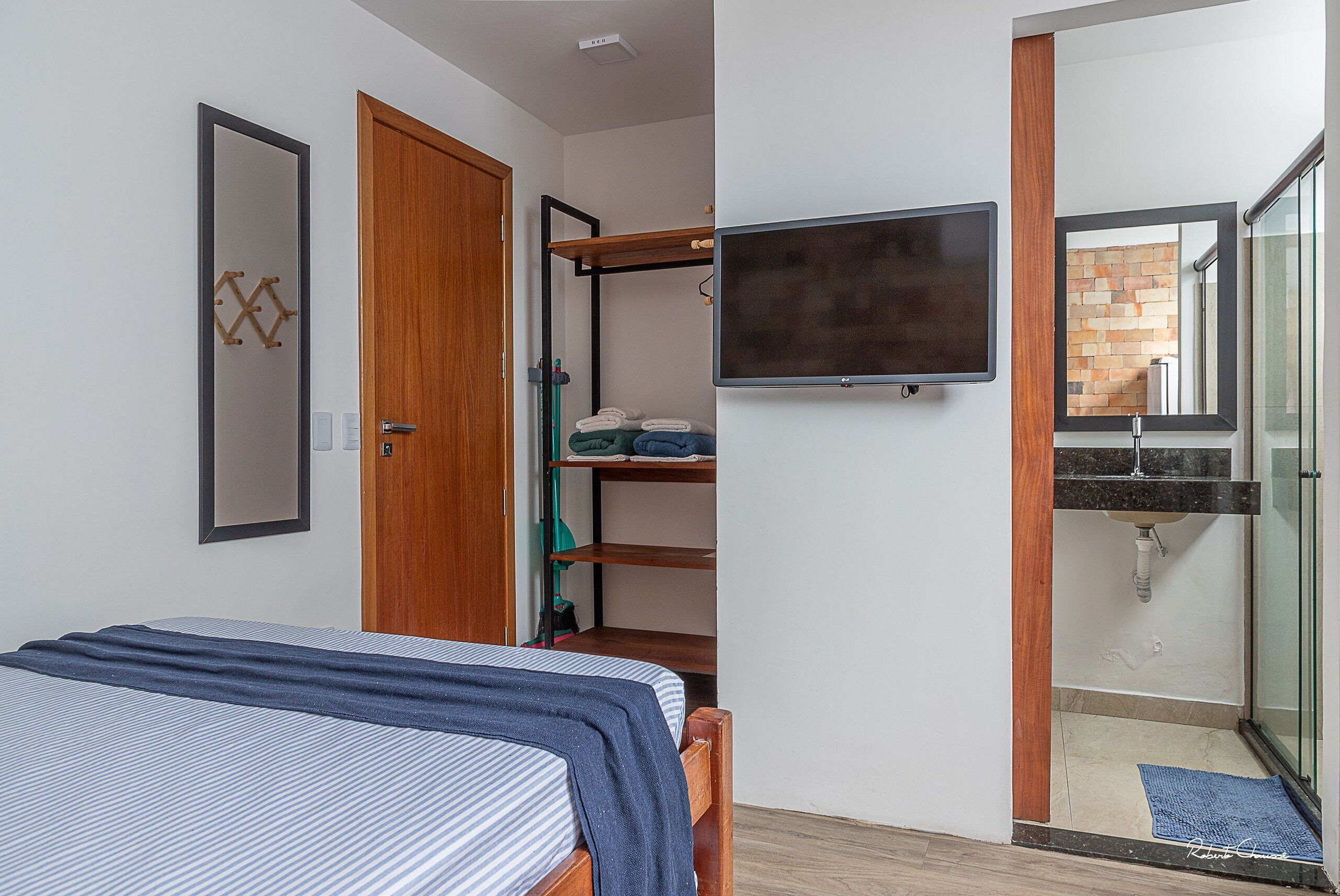 Un dormitorio con cama, armario, televisor montado en la pared y baño con ducha y lavabo.