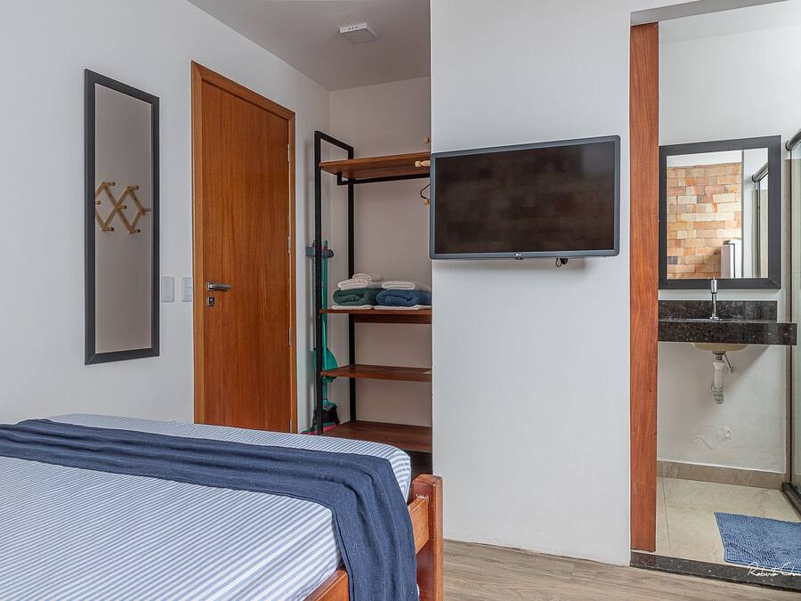 Un dormitorio con cama, armario, televisor montado en la pared y baño con ducha y lavabo.