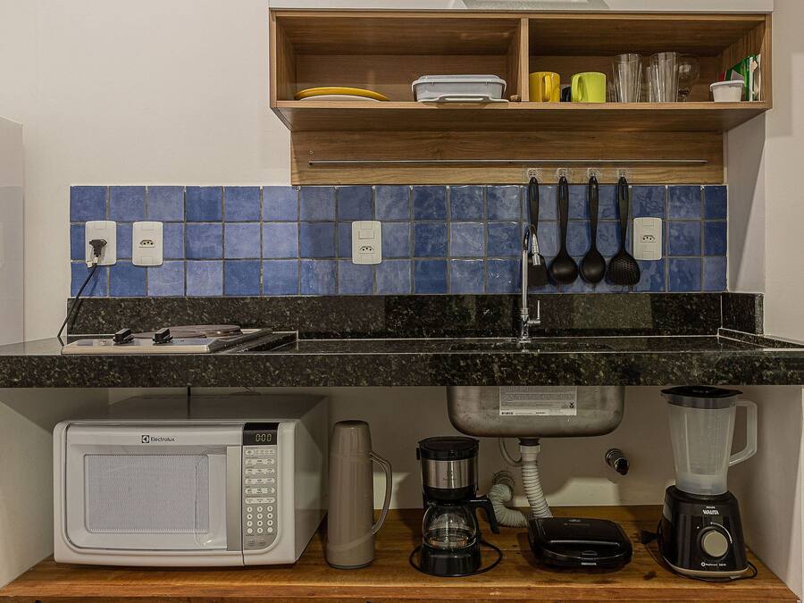 A kitchen with a microwave, coffee maker, and blender on the counter, a wooden shelf above with dishes, and blue tiles on the backsplash.