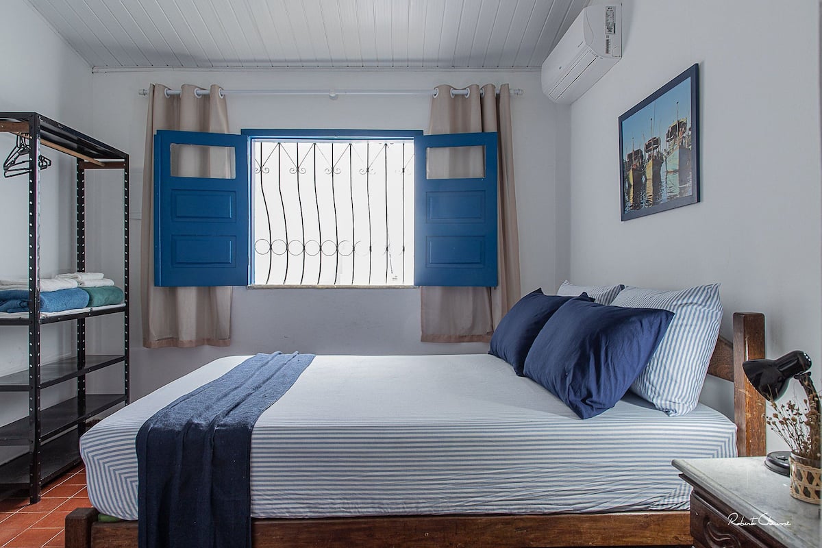 Ein Schlafzimmer mit einem Bett, einem Fenster mit blauen Rollläden, einer Hängelampe und einem Wandbild.