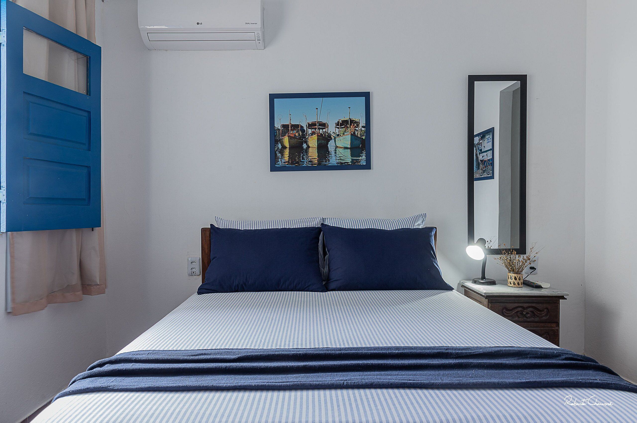 Una cama bien hecha con ropa de cama azul, una mesita de noche con una lámpara, un cuadro enmarcado en la pared y una puerta azul.