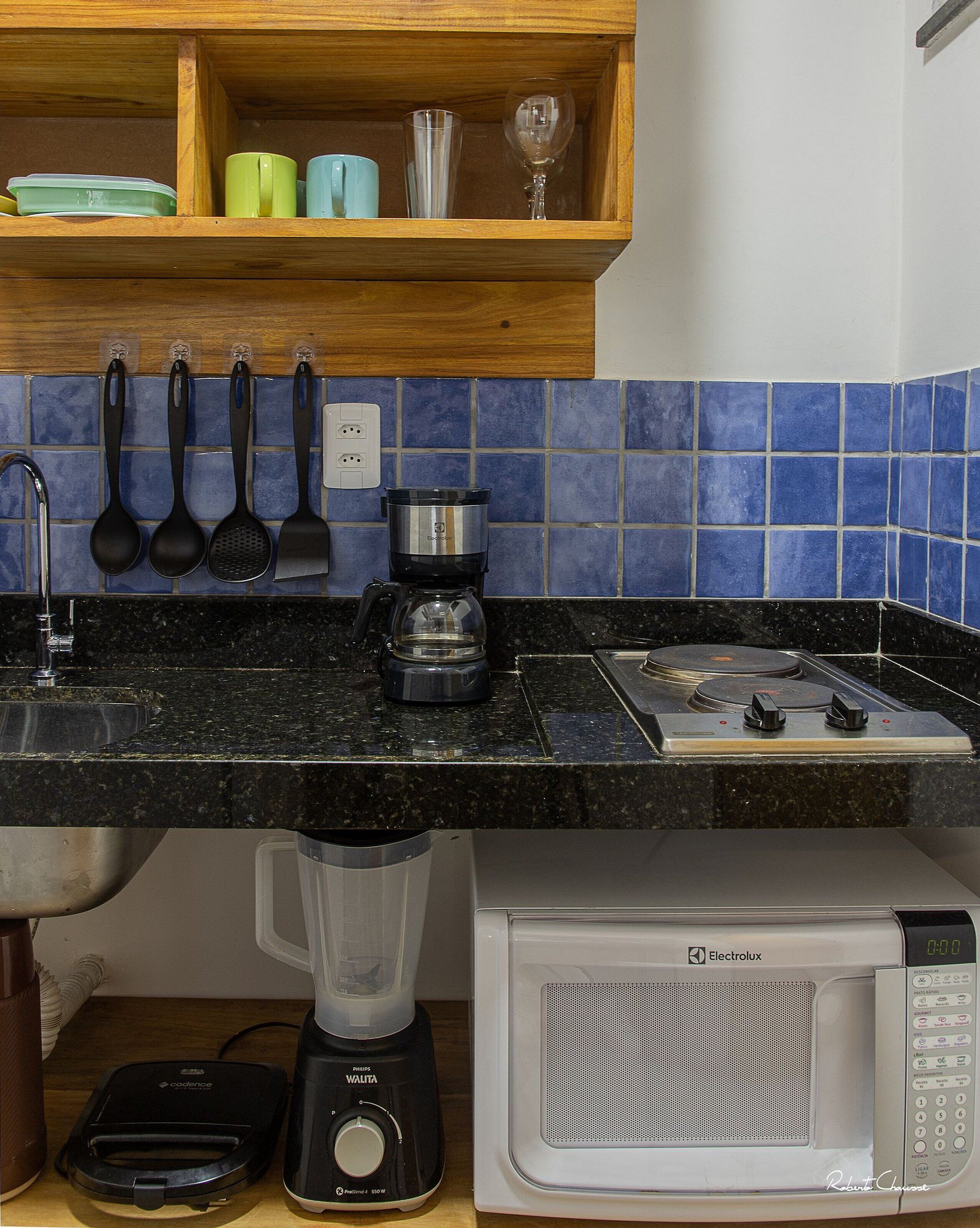 Ett kök med en svart bänkskiva, en kaffebryggare, en mixer, en mikrovågsugn och en hylla med koppar och ett vinglas.