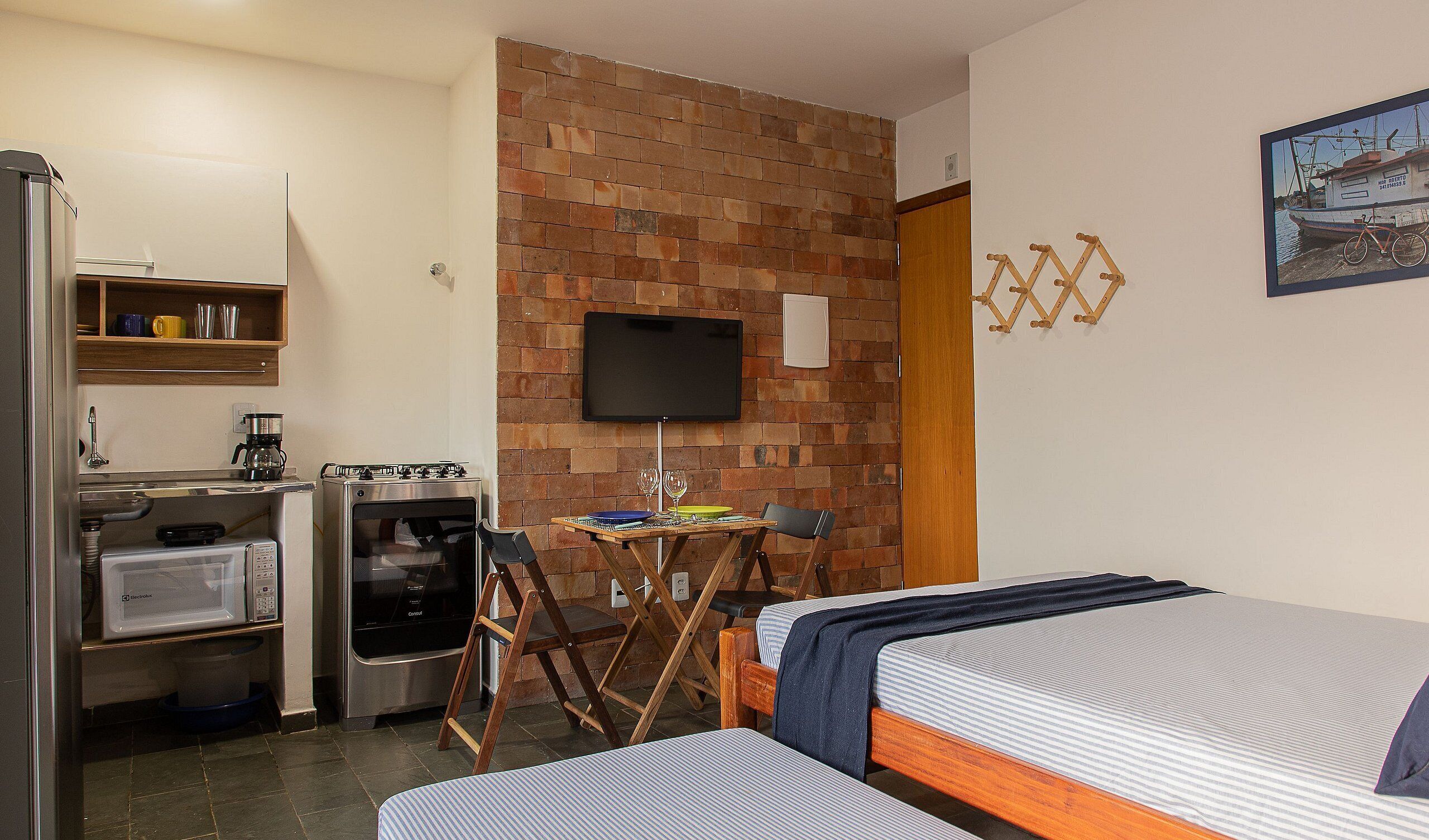 A compact room with a kitchenette, a TV mounted on a brick wall, and a bed with a striped bedspread.