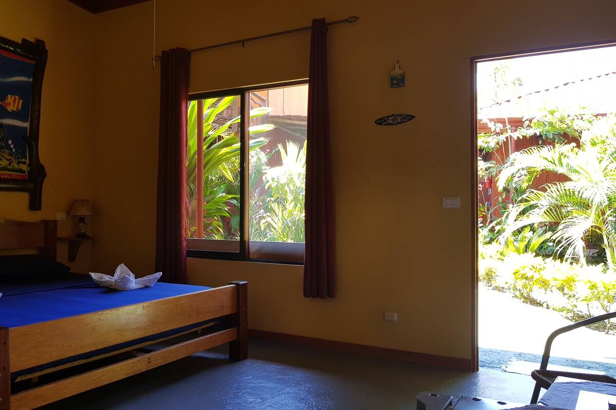 Quarto com cama, janela com cortinas, um quadro na parede e vista para o verde lá fora.