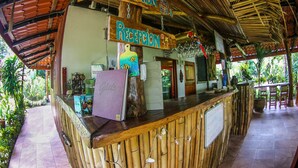 Zona de recepción con mostrador de bambú, un cartel que indica 'Reception' y 'Bebidas', y vistas al jardín exterior.
