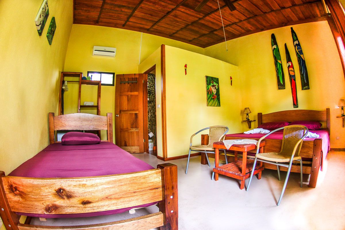 A room with a wooden bed, a small table with chairs, a wooden wardrobe, and a door leading to another room.