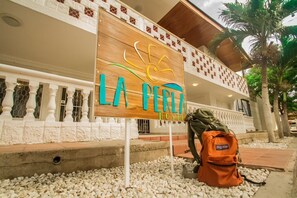 A sign for "LA PERLA HOSTEL" with a sun graphic, set against a building with a white facade and a balcony. - La Perla Hostel (Santa Marta)