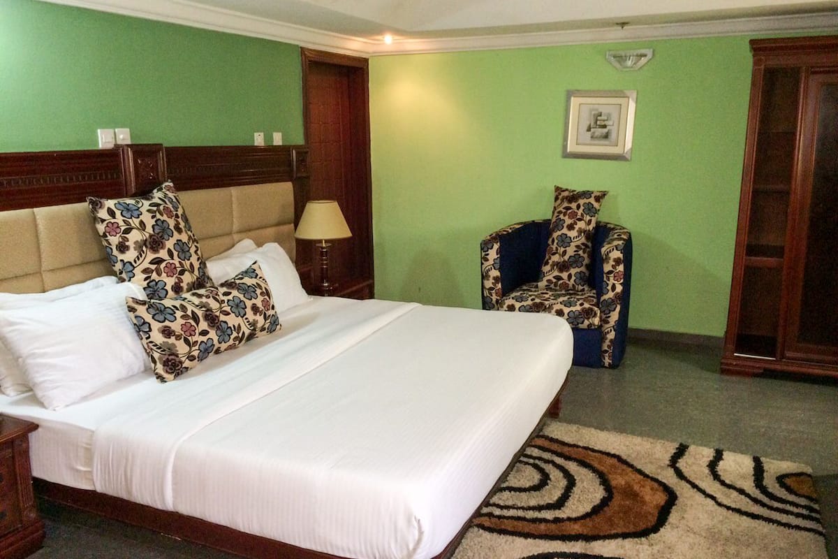 A hotel room with a large bed, floral-patterned pillows, a nightstand, a lamp, a chair, and a carpet.
