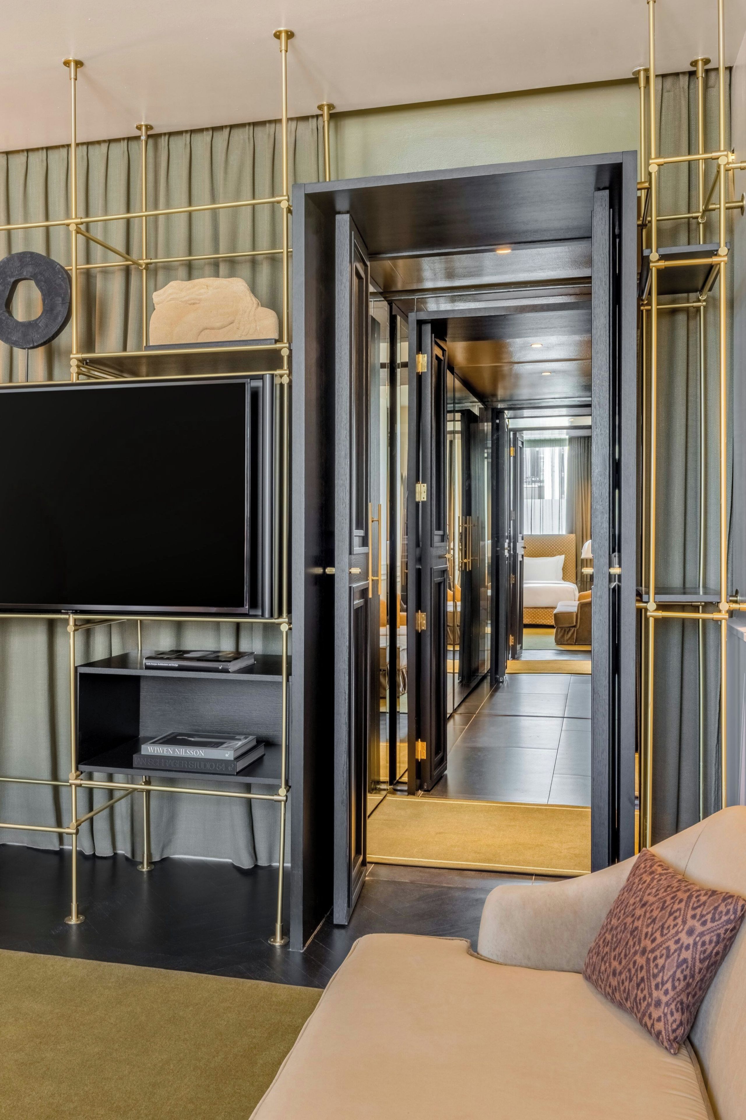 Ein modernes Wohnzimmer mit einem Flachbildfernseher an einer schwarzen Wandhalterung, einer Couch mit einem gemusterten Kissen und einem Bücherregal mit Büchern und Dekorationsobjekten.