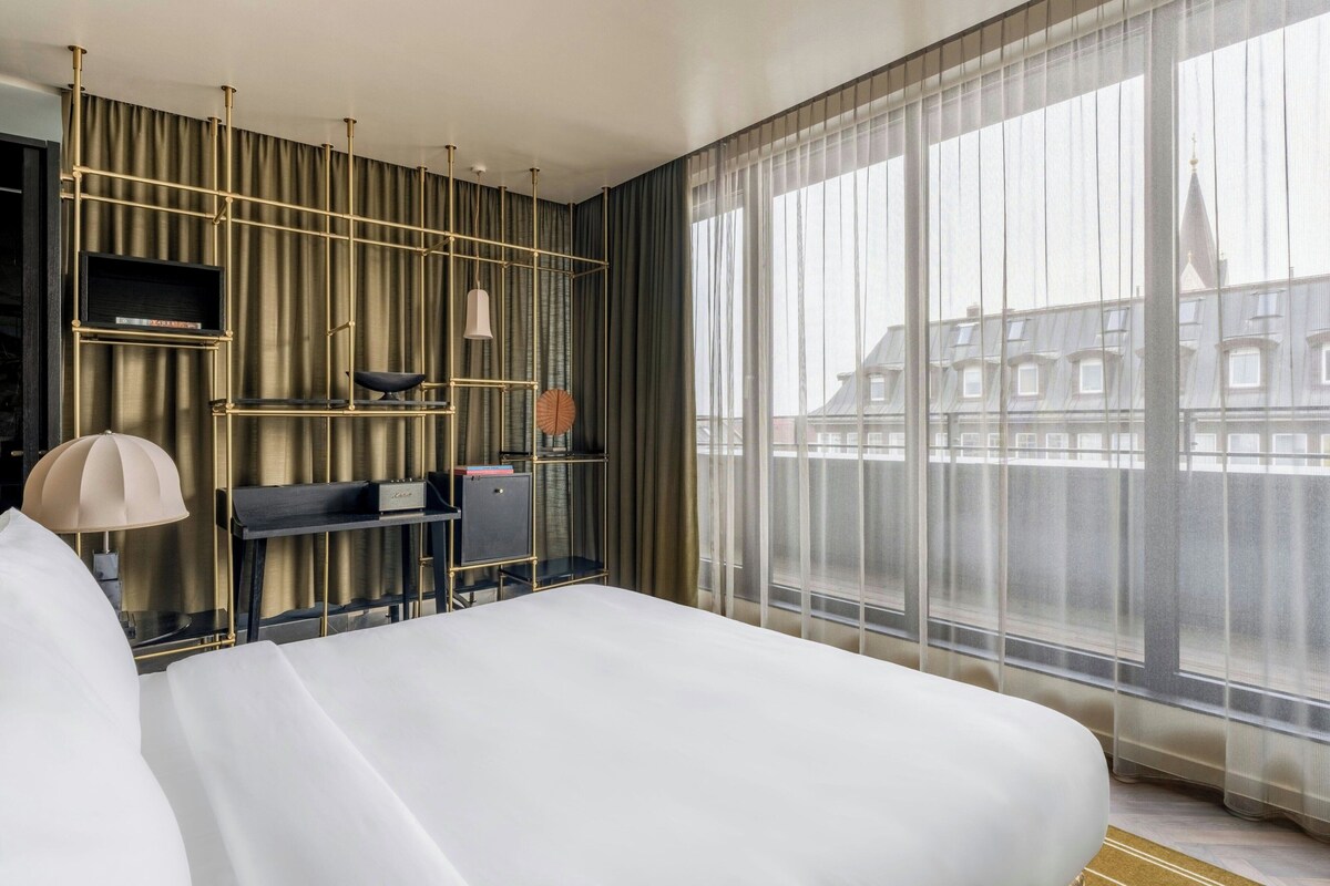 A hotel room with a large bed, a desk with a chair, a TV, and a view of buildings through sheer curtains.