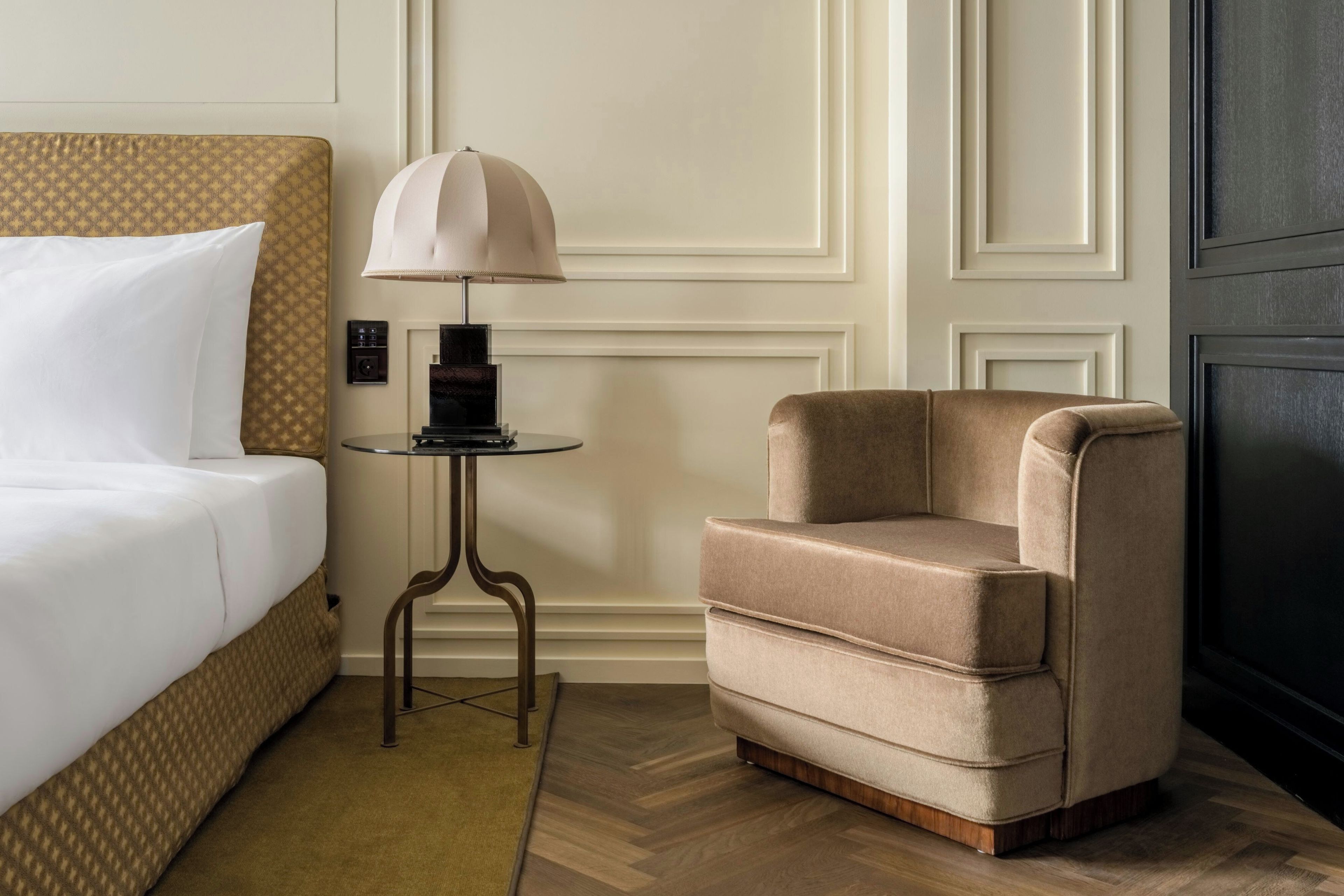 Una camera d'albergo con un letto, un comodino con una lampada, una sedia e un pavimento in legno.