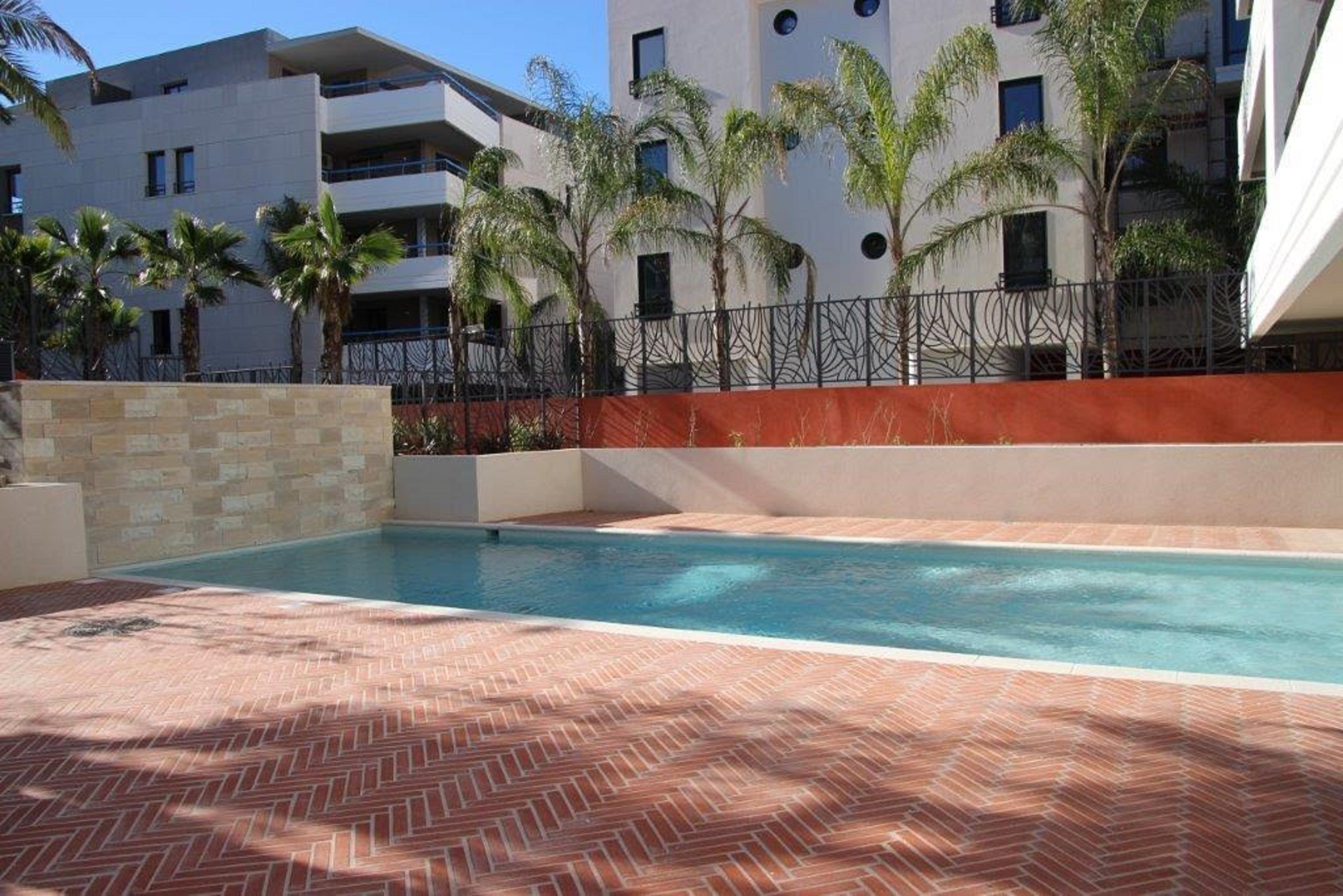 Une piscine avec une terrasse en briques, entourée d’une basse clôture et de palmiers, et un bâtiment moderne en arrière-plan.