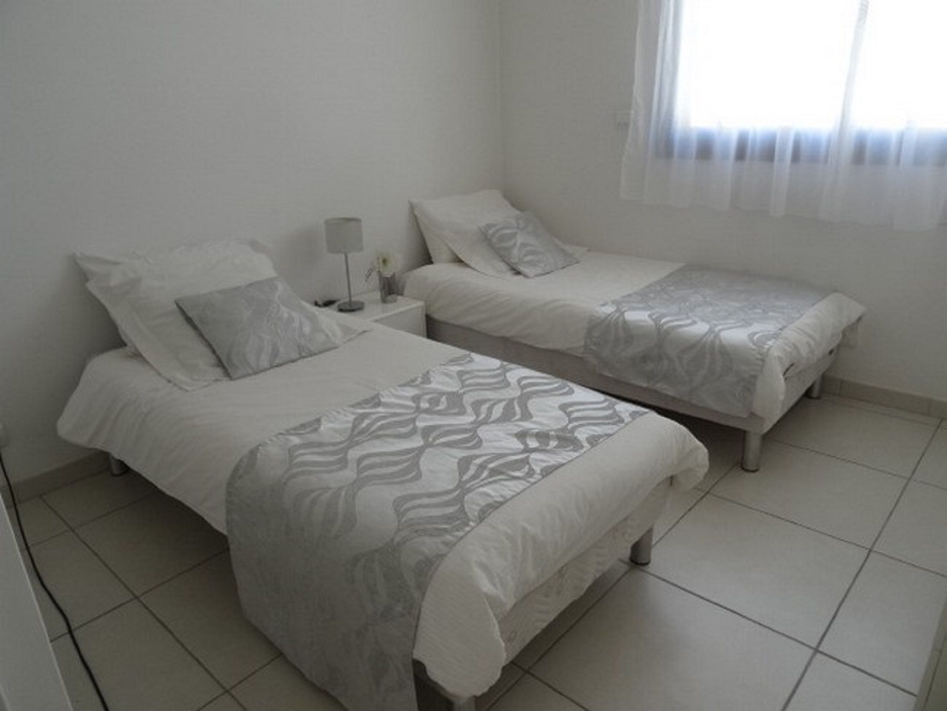 A bedroom with two beds, a nightstand, a lamp, and a window with sheer curtains.