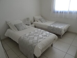A bedroom with two beds, a nightstand, a lamp, and a window with sheer curtains.