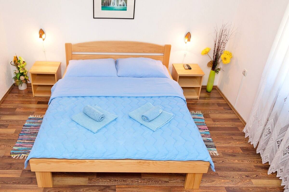A neatly made bed with a blue quilt and two rolled towels, wooden headboard, bedside tables with lamps, a framed picture on the wall, and a vase with flowers.