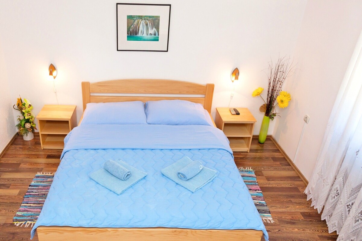 A neatly made bed with a blue quilt and two rolled towels, wooden headboard, bedside tables with lamps, a framed picture on the wall, and a vase with flowers.
