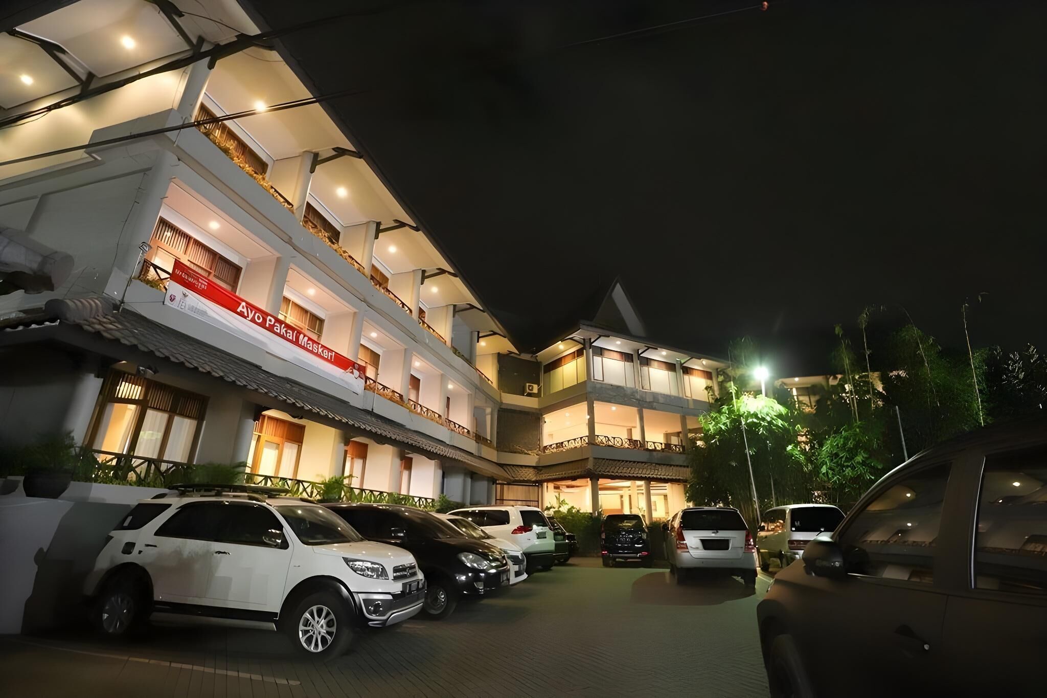 A multi-story hotel with a balcony on the upper floor, parked cars, and a sign indicating a mask requirement.