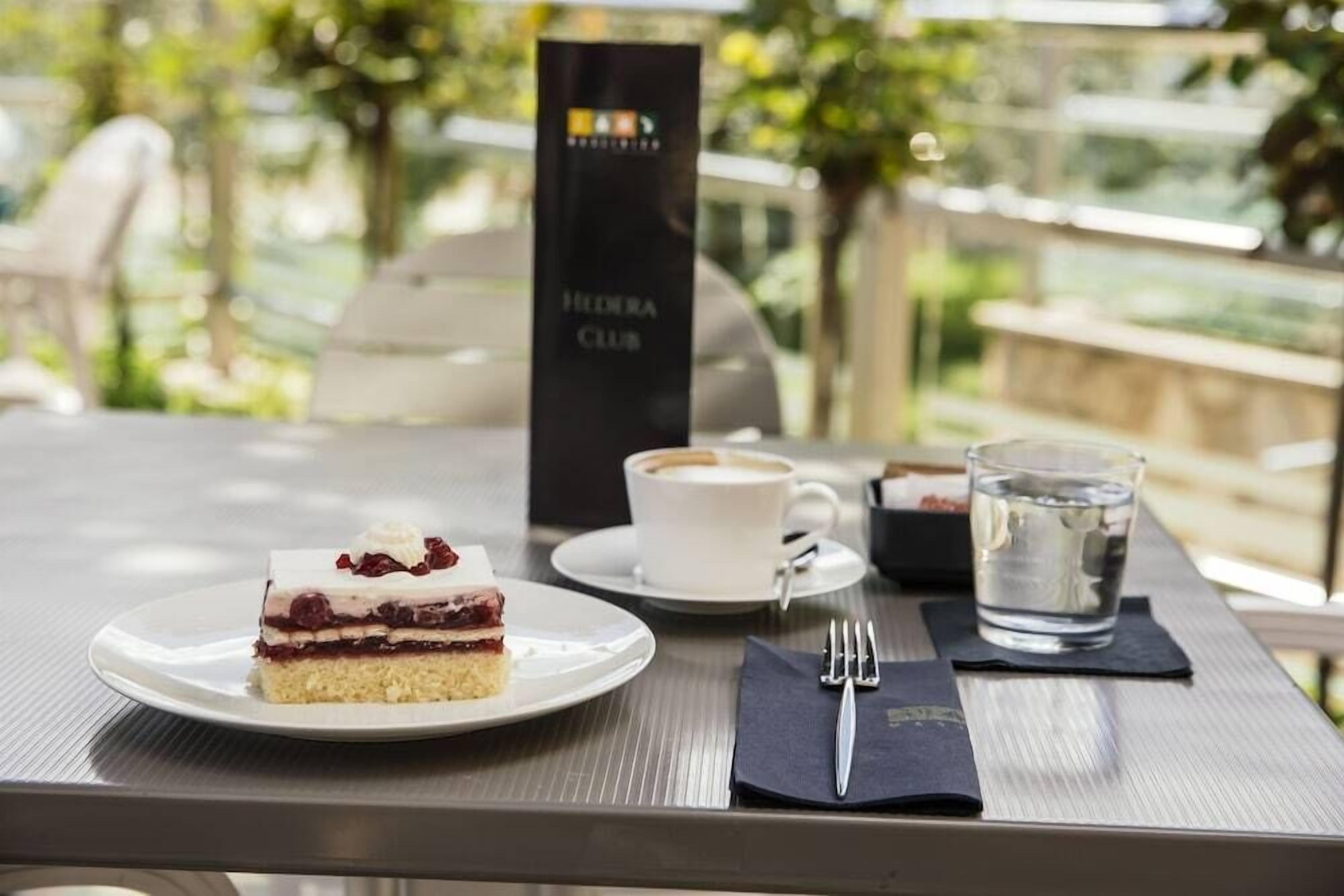 A table with a slice of cake, a cup of coffee, and a glass of water on a terrace with a view of greenery.