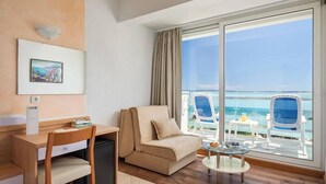A hotel room with a sofa, a desk, a chair, a small table, and a view of the ocean through large windows.