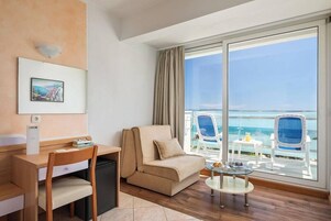 Quarto de hotel com sofá, escrivaninha, cadeira, mesinha e vista para o mar através de grandes janelas.