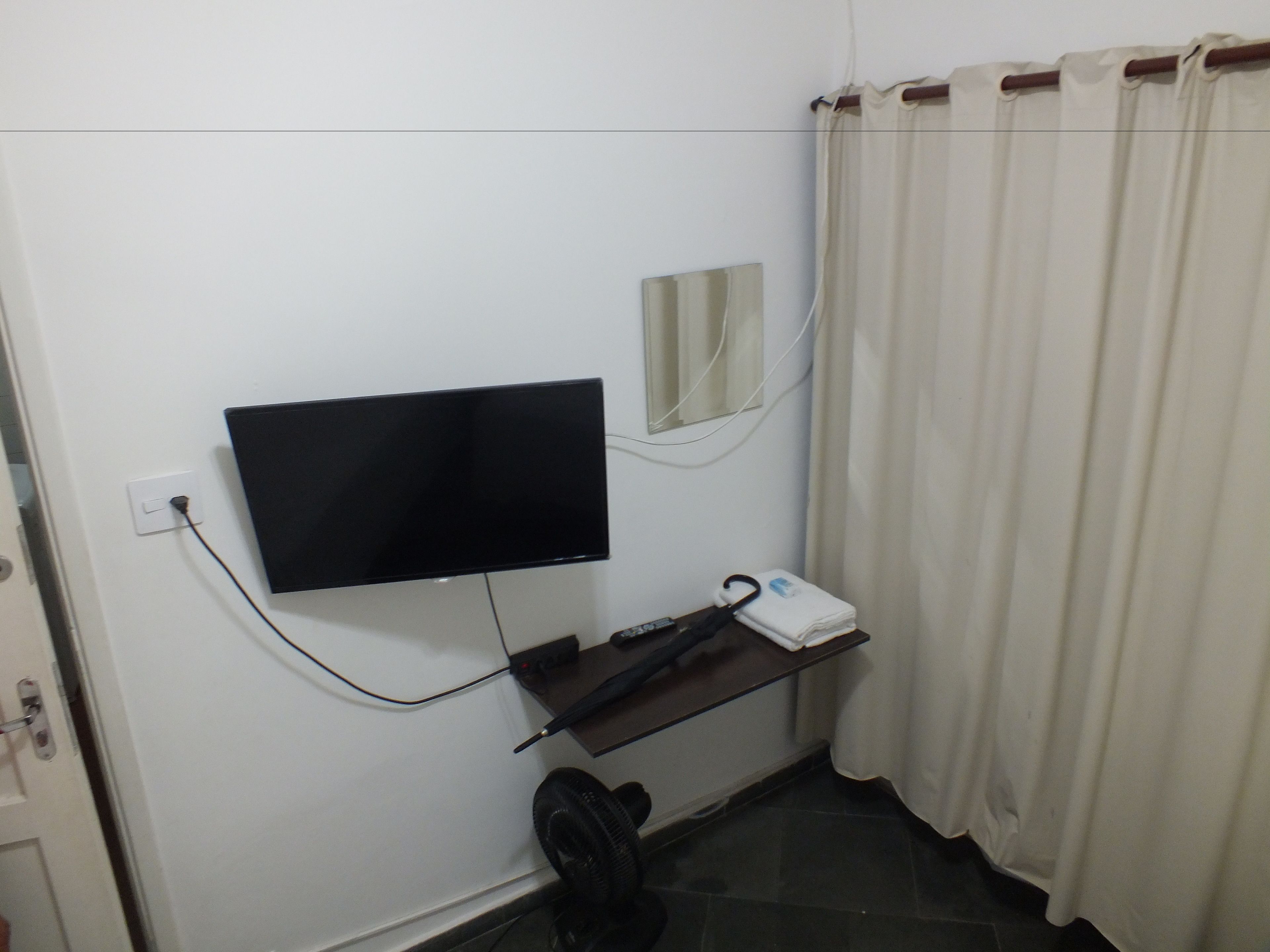 A room with a TV mounted on the wall, a small shelf with a remote control, and a window with curtains.