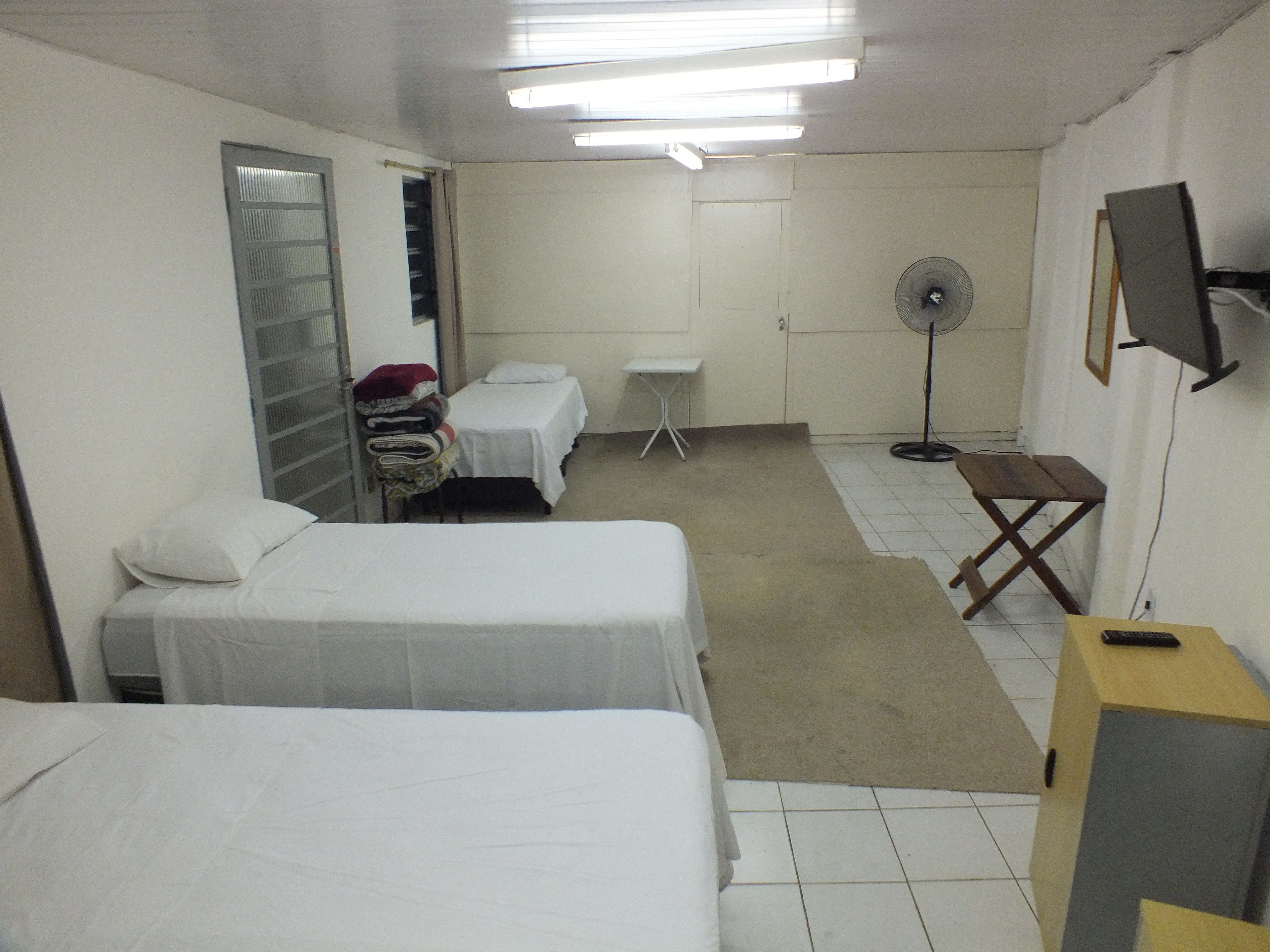 Quarto de hotel com duas camas de solteiro, uma mesa pequena, um ventilador e uma TV sobre um suporte.