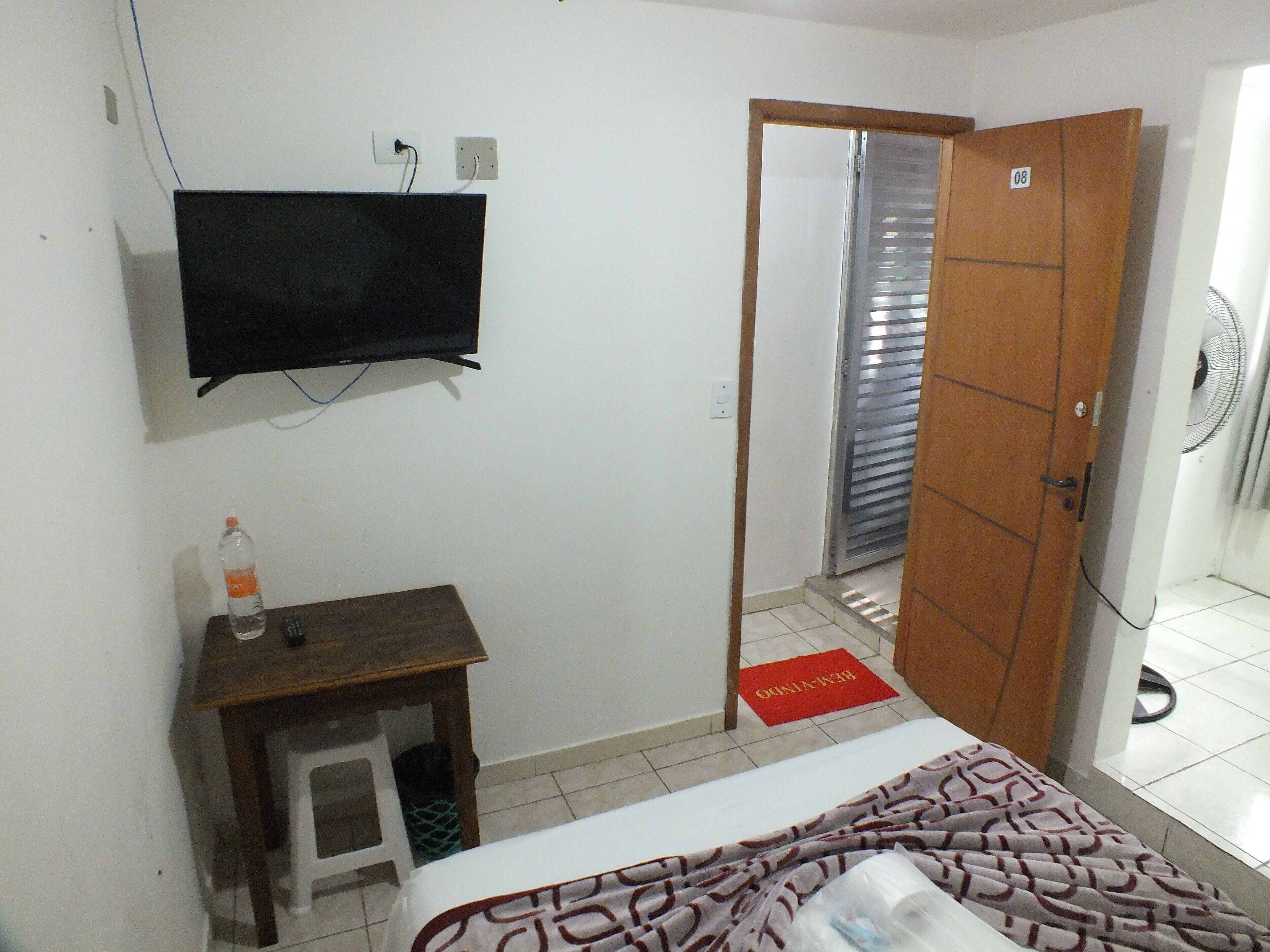 A hotel room with a bed, a TV mounted on the wall, a small wooden table, a water bottle, and a red doormat.