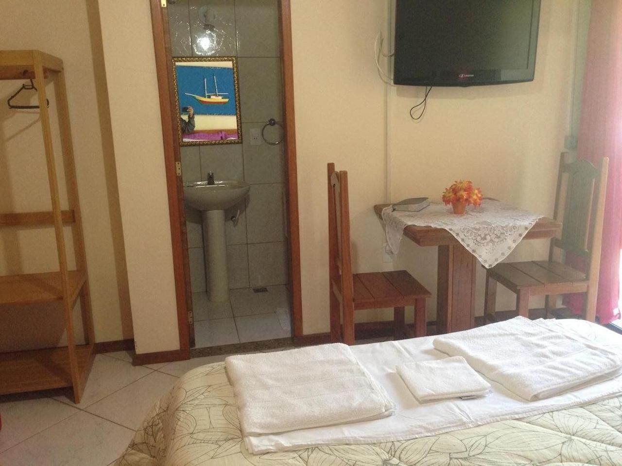 Une chambre d’hôtel avec un lit, une petite table sur laquelle se trouve un vase, une chaise en bois, un téléviseur fixé au mur et une salle de bain visible à travers une porte ouverte.