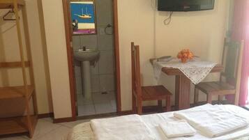 Une chambre d’hôtel avec un lit, une petite table sur laquelle se trouve un vase, une chaise en bois, un téléviseur fixé au mur et une salle de bain visible à travers une porte ouverte.