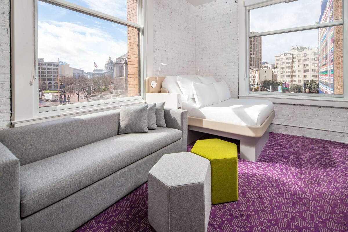 A modern room with a grey sofa, a white bed, and a yellow ottoman.