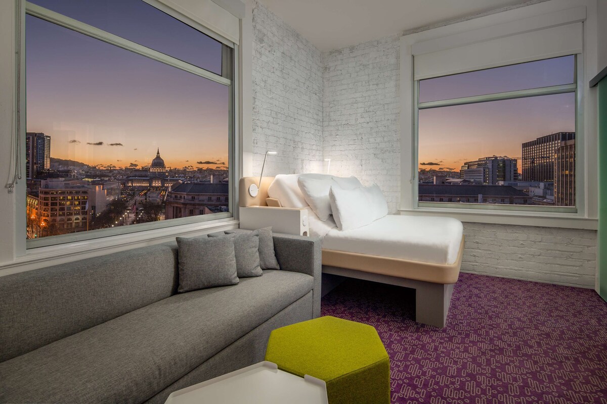A modern hotel room with a city view, a grey sofa, a bed, and a small green ottoman.