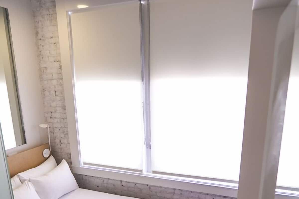 A modern hotel room with a large bed, a bedside table, a window with blinds, and a brick wall.