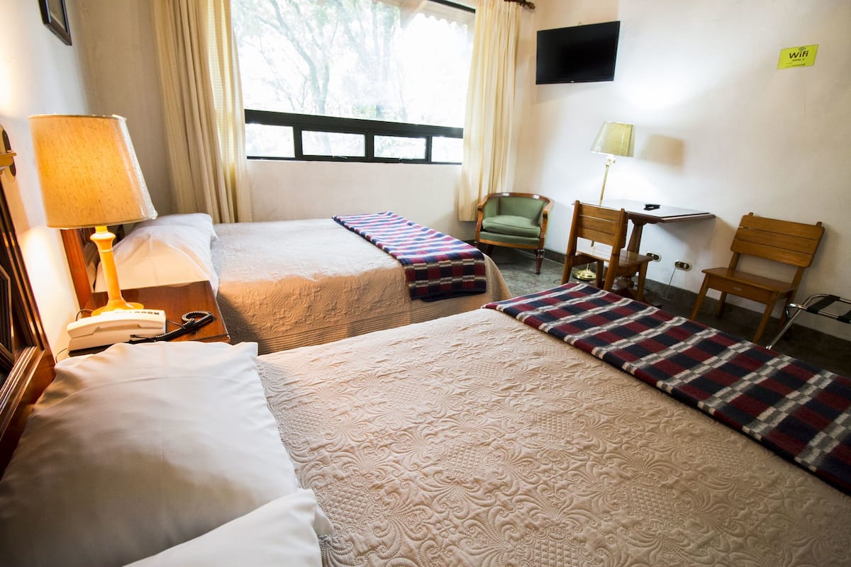 A hotel room with two beds, a desk, a chair, a lamp, and a TV on the wall.