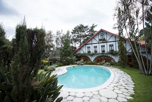 Una piscina con borde de piedra, rodeada de follaje y un edificio de fachada blanca y tejado rojo.