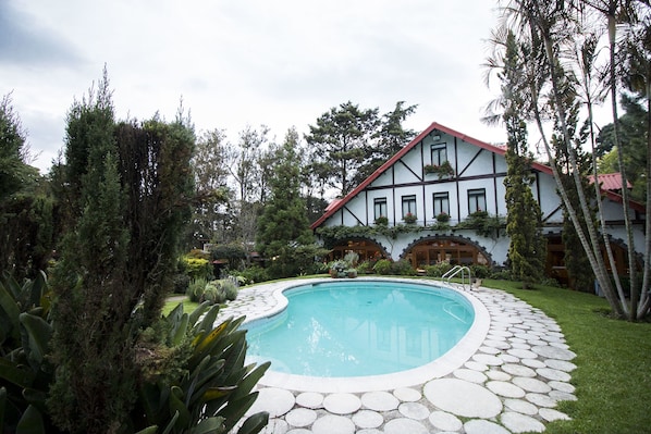 A pool with a stone border, surrounded by greenery and a building with a white facade and red roof.
