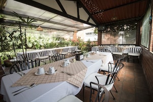 Un área de comedor al aire libre con mesas dispuestas para una comida, rodeada de sillas y vistas a un jardín.