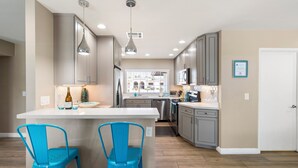 Une cuisine moderne avec des meubles gris, un îlot central blanc avec deux chaises bleues et une grande fenêtre donnant sur des immeubles.