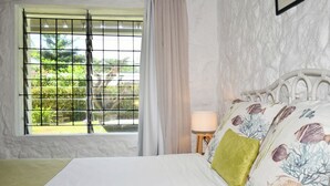 Un dormitorio con cama, ventana con vistas a follaje, lámpara y pared con textura.