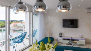 Een moderne woonkamer met een grote televisie, een glazen salontafel en een bank. Er is een balkon met uitzicht op het water en de gebouwen.