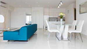 Een moderne woonkamer met een blauwgroene bank, een glazen eettafel met witte stoelen en een keuken op de achtergrond.