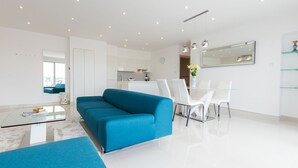 Een moderne woonkamer met een blauwe bank, een glazen salontafel, en een eethoek met witte stoelen en een glazen tafel.