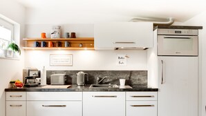 Una cocina moderna con armarios blancos, horno empotrado, cafetera y hervidor. Hay estantes con tazas y un letrero que dice "Butter bei der Fischerei".