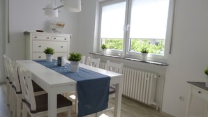 Zona pranzo con tavolo bianco, tovaglia blu e piante in vaso. Si vede una finestra con persiane, un termosifone bianco e un mobiletto bianco con cassetti.