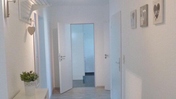 Un corridoio con pavimento in legno, pareti bianche e una porta in fondo. Sul lato sinistro, un piccolo vaso con una pianta su un tavolo bianco.