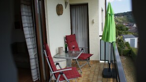 A balcony with a table and two red chairs, a green umbrella, and a view of a hillside. - Wonderful view of the Nahe Valley and the Frombork (Sonnenberg-Winnenberg)
